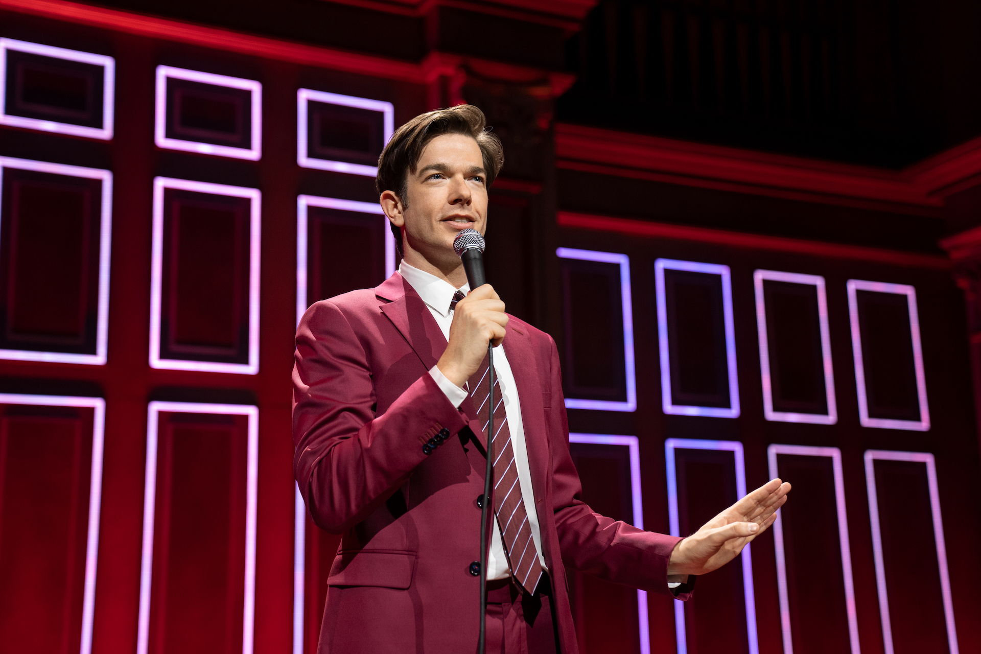 Plan moyen du comédien John Mulaney en costume et cravate bordeaux, debout devant un fond de scène recouvert de rectangles de néon ;  toujours de son spécial "Bébé J"